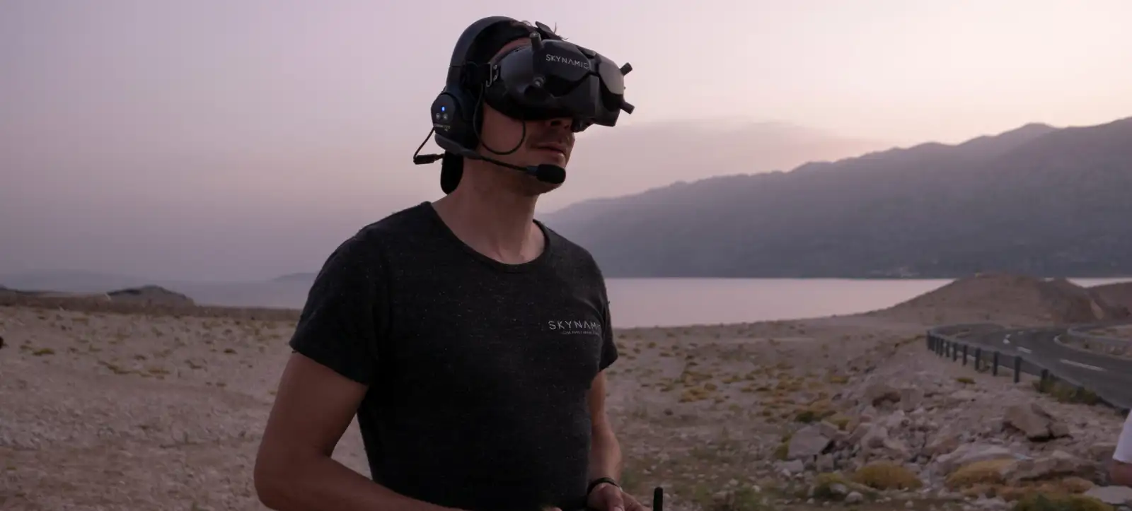 Skynamic FPV-Pilot mit Goggles und Headset, Controller in den Händen, an der Küste bei Dämmerung